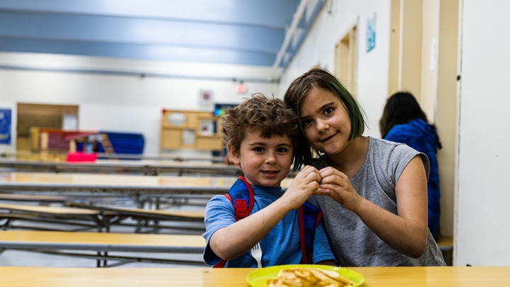 Two young children sit side by side at a table in a school cafeteria, holding a small object together and facing the camera. One child wears a blue shirt with red backpack straps, and the other has short hair with green highlights and wears a grey shirt. A plate with food is visible in the foreground, while cafeteria tables and another person seated in the background appear slightly out of focus.