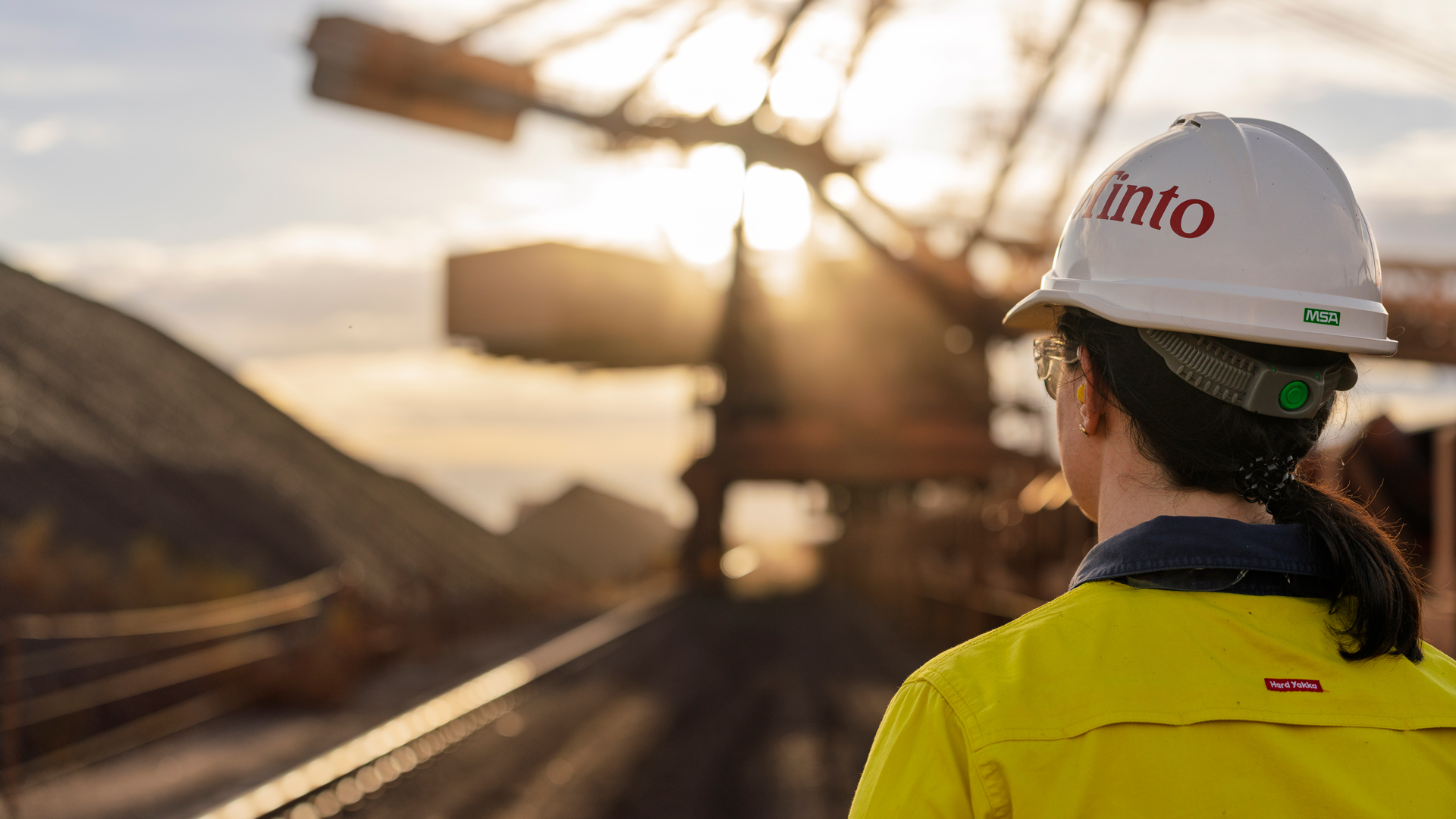 Person wearing a white hard hat and high-visibility shirt standing beside industrial rail tracks, with large machinery and a setting sun in the background.