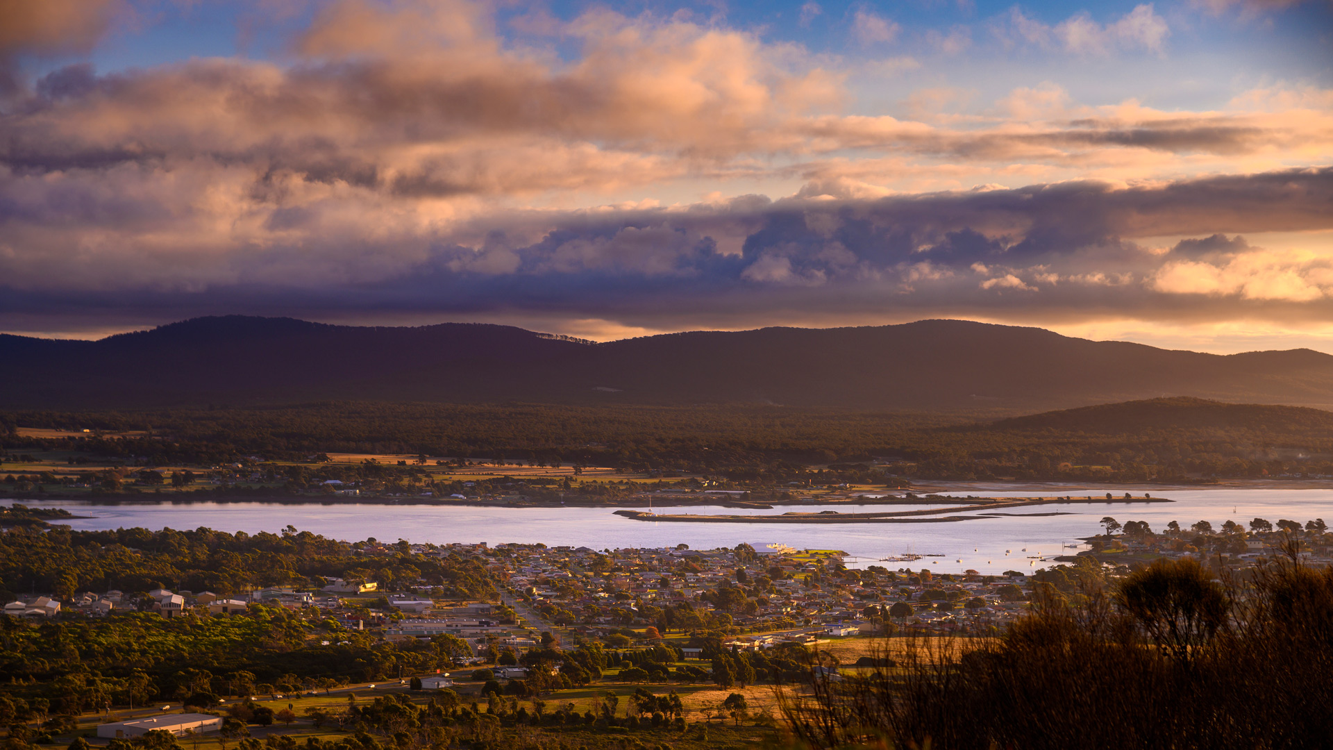 A scenic view of a coastal town at sunset, with a river or estuary in the middle ground reflecting soft golden light. 