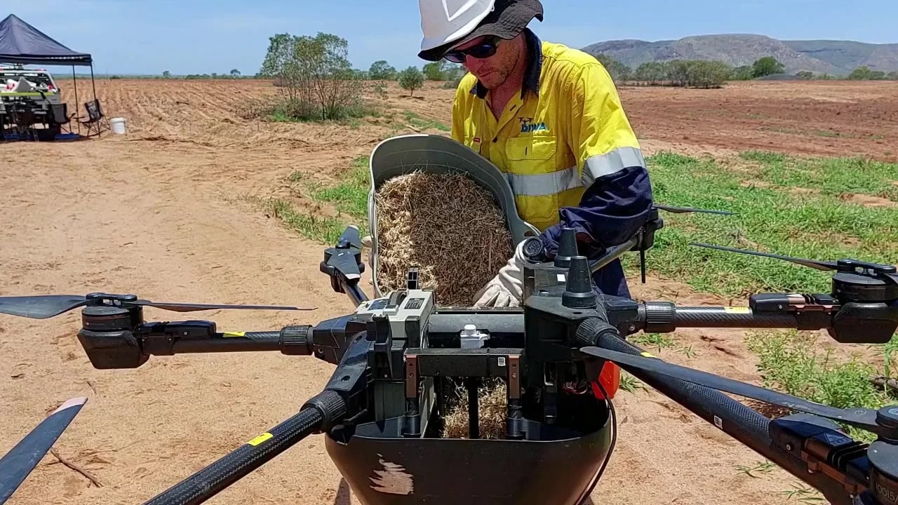 A worker wearing high visibility gear poring seeds into a basket attached to a drone near a farmland. 