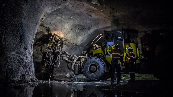 Yellow underground mining vehicle with a large drill attachment parked in a dimly lit rock tunnel, with two workers in high-visibility clothing beside it and water reflecting the equipment below.