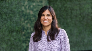A person with long dark hair smiles at the camera while standing outdoors in front of a green, leafy hedge, wearing a patterned button-up shirt.