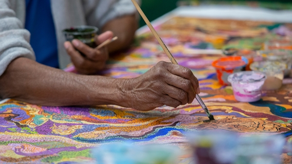 A close-up of a hand holding a paintbrush, painting a vibrant artwork with intricate patterns in purple, orange, yellow, blue, and green. 