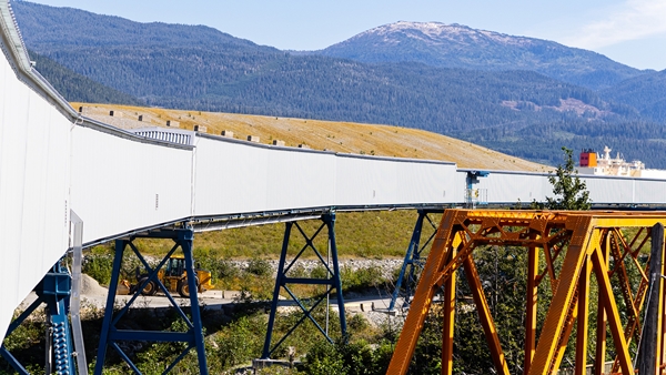 Elevated industrial conveyor belt supported by steel structures, with forested mountains in the background.