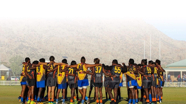 Australian rules football players huddle together on a football field, with goal posts and rugged hills in the background.
