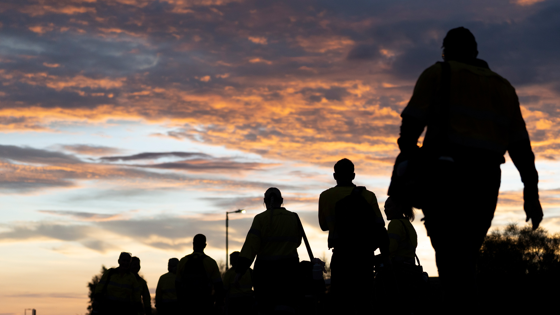 Silhouettes of several people wearing high-visibility work clothing walk together outdoors at dawn or dusk, carrying bags and equipment against a sky filled with orange and purple clouds, with a streetlight visible in the background.