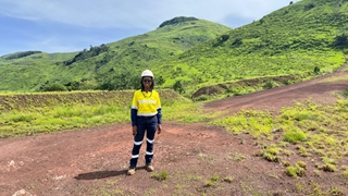 A person wearing a high-visibility work uniform, hard hat, and safety glasses stands on a reddish dirt road in a lush, green hilly landscape, with rolling hills and a cloudy sky in the background.