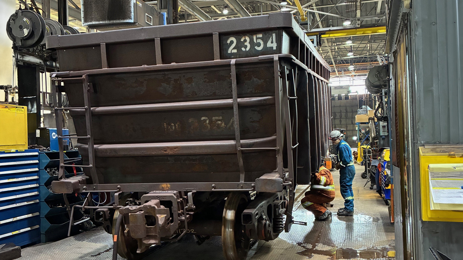 Railcar labeled “2354” inside an industrial workshop, with two workers conducting maintenance near the wheel assembly, surrounded by tools and equipment on a factory floor.