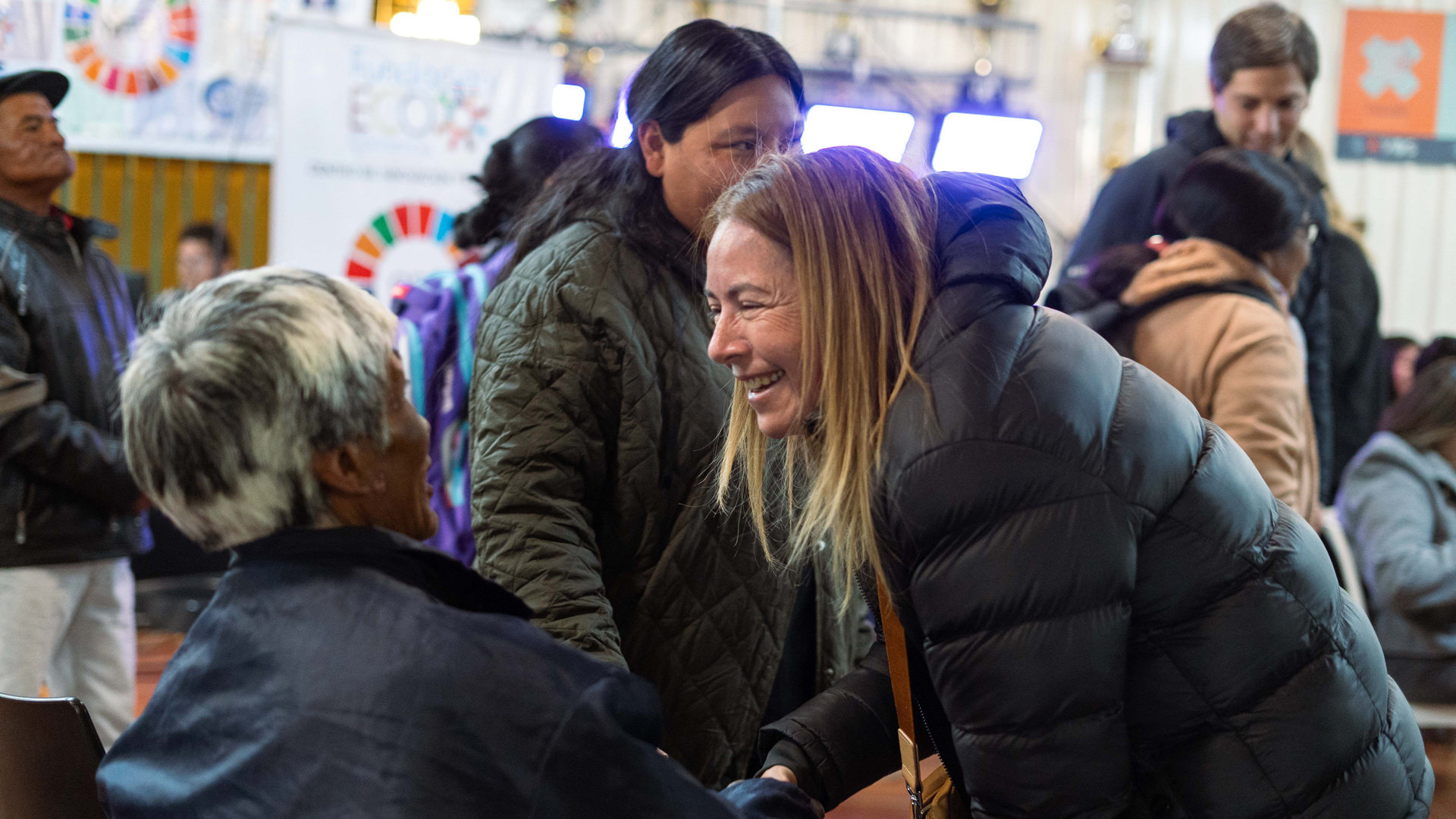 Las personas se saludan y conversan en una reunión comunitaria en un espacio interior, con participantes sentados y de pie entre paneles informativos y señalización del evento al fondo.