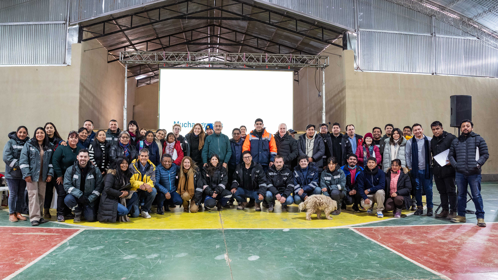 Foto grupal grande de aproximadamente 40 personas reunidas dentro de un polideportivo cubierto, posando en una cancha de baloncesto frente a una pantalla de proyección y una estructura de escenario. Muchos participantes llevan chaquetas de trabajo y ropa para actividades al aire libre, y un perro se encuentra cerca de la primera fila.