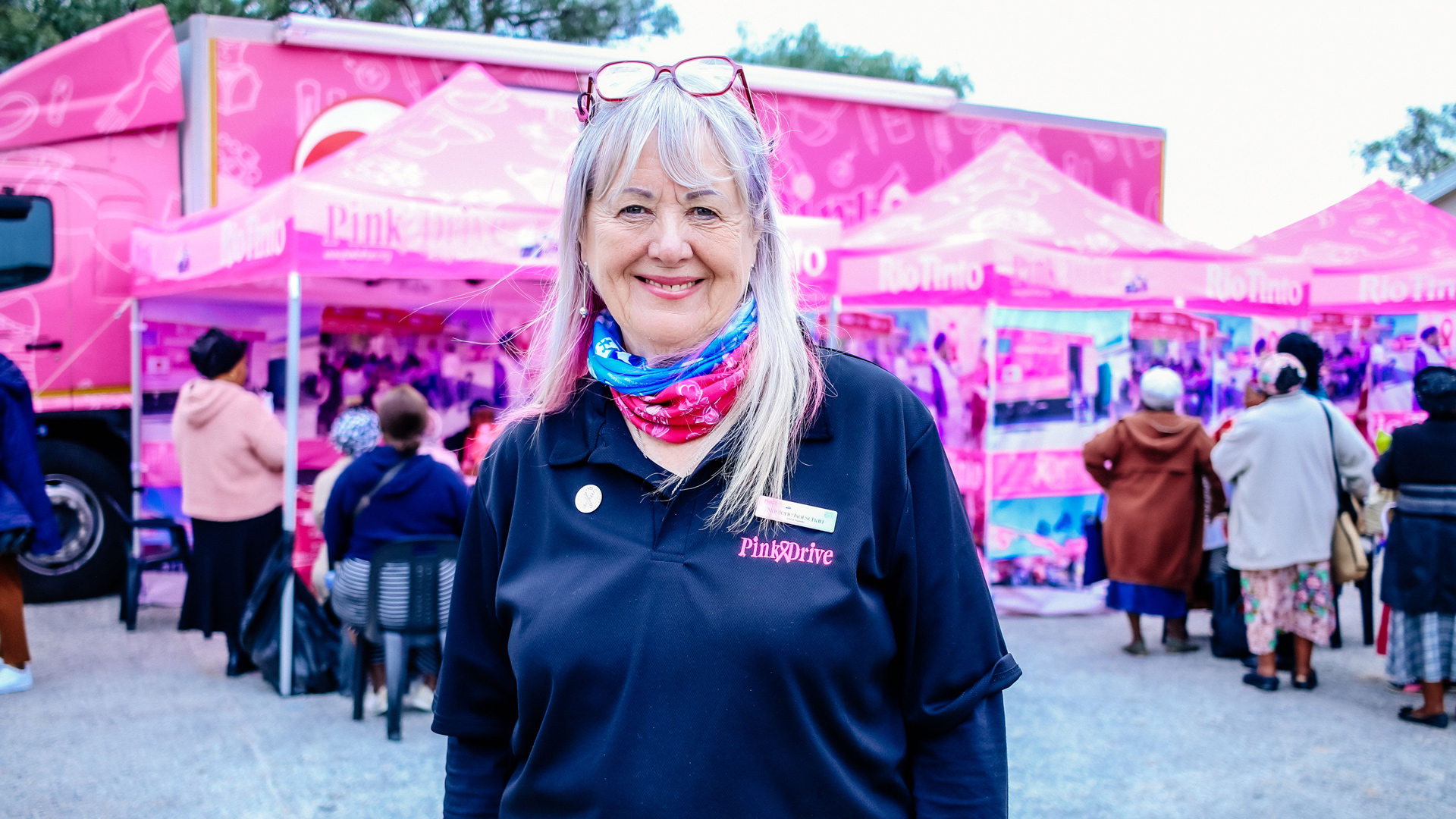 Noelene Kotschan, CEO and Founder of PinkDrive NPC posing for the camera in front of a community stall setting with pink tents and a truck.