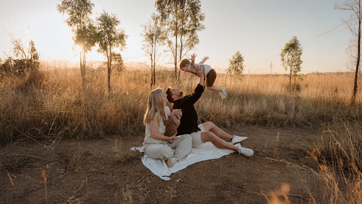 Renee and Chris at an outdoor picnic; Chris joyfully lifting baby into the air, while Renee on side holding another baby, surrounded by trees.