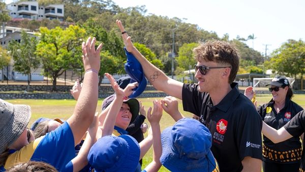 A group of children wearing blue and yellow sports uniforms and hats reach up for high-fives with an adult coach on a sunny outdoor sports field.