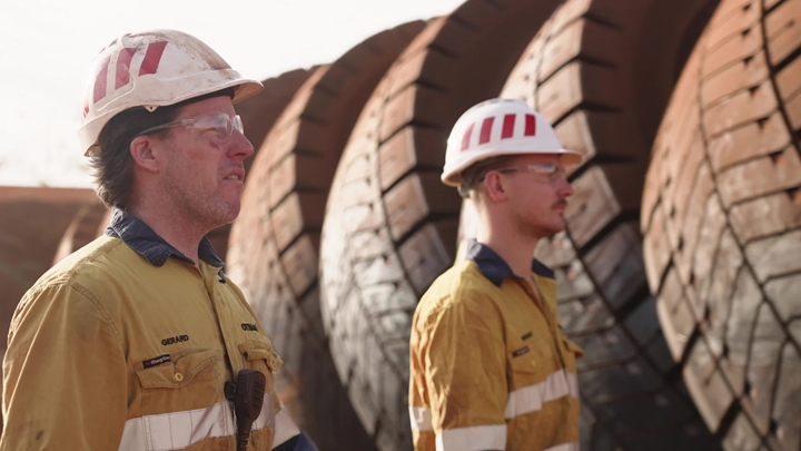 Two 91������ employees wearing a safety hat and facing ahead in front of large mine truck tyres. 