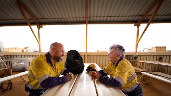 Two workers wearing yellow high-visibility shirts sit across from each other at a long table inside a covered outdoor structure.