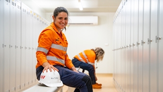Taylee, a production supervisor at Rio Tinto’s Amrun site, sitting on a bench in a locker room wearing high-visibility orange workwear with reflective strips. 