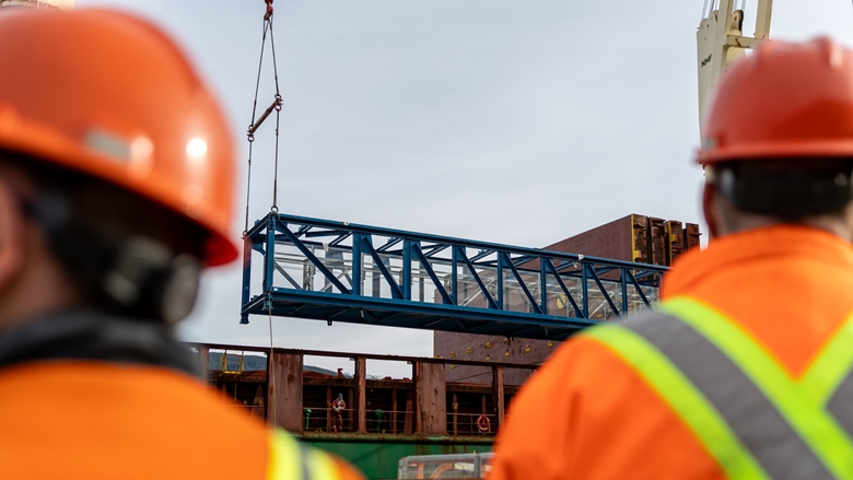 Industrial workers in high‑visibility protective clothing watch as a blue steel conveyor truss is lifted by cranes at an industrial site, with buildings and equipment visible in the background under a clear sky