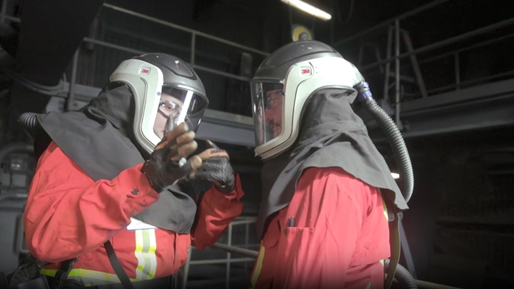 Two individuals in red protective suits and helmets with face shields and attached hoses, standing in an industrial setting with metal railings and equipment.