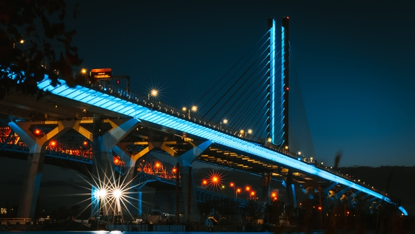 A cable-stayed bridge crosses the scene at night, with a tall illuminated pylon and blue-lit roadway standing out among streetlights and urban infrastructure.