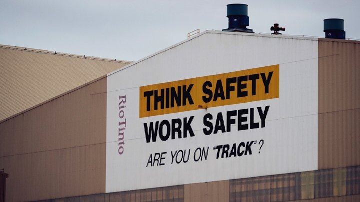 Industrial building with a large safety message painted on the side reading 'THINK SAFETY WORK SAFELY ARE YOU ON 'TRACK'?', with 'THINK SAFETY' highlighted in yellow. The building features two blue exhaust stacks and a vertical 'Rio Tinto' label on the left side.