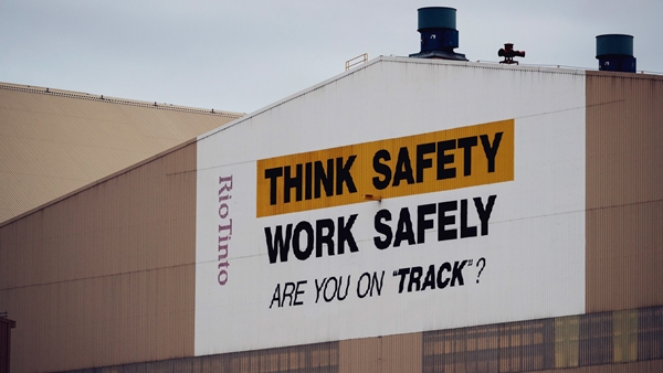 Industrial building with a large safety message painted on the side reading 'THINK SAFETY WORK SAFELY ARE YOU ON 'TRACK'?', with 'THINK SAFETY' highlighted in yellow. The building features two blue exhaust stacks and a vertical 'Rio Tinto' label on the left side.