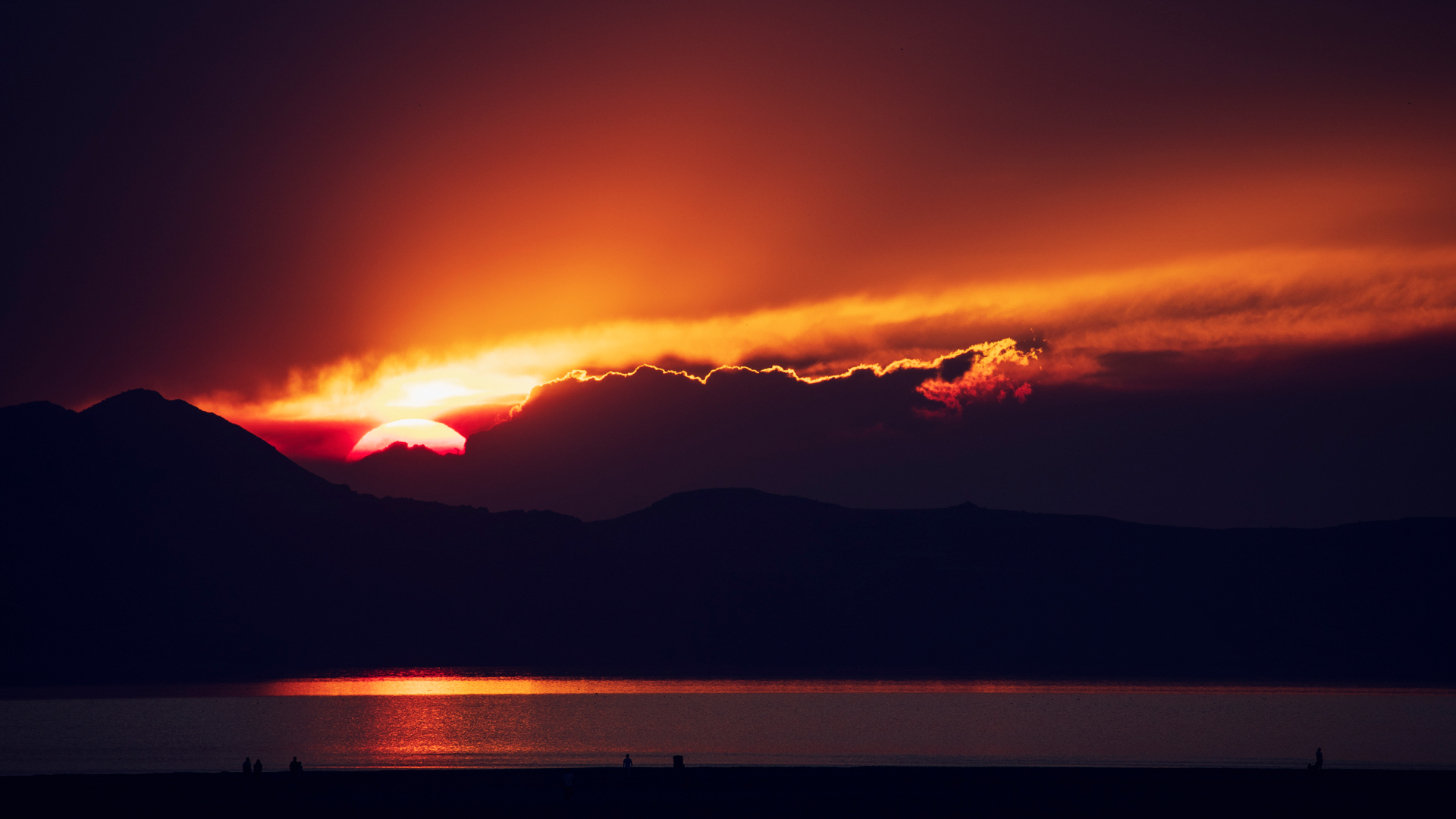 Sun setting behind dark mountain silhouettes, with glowing orange clouds and a reflection across calm water.