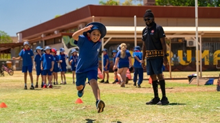 A child wearing a blue athletic outfit and sun hat runs and jumps over a rope obstacle on a grassy field, while a group of similarly dressed children and an adult stand in the background near a school building during an outdoor sports activity.