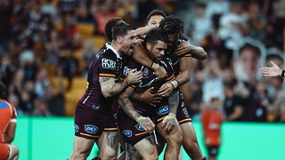 Four football players in maroon uniforms huddle and embrace in celebration on a stadium field, with a blurred crowd in the background and visible sponsor logos on their jerseys.