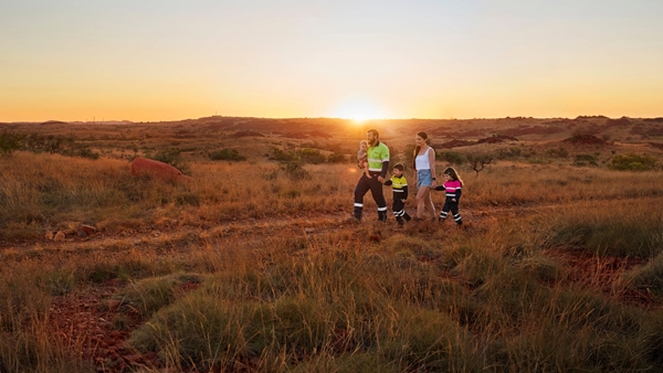 A group of adults and children walk through dry grassland at sunset, with rolling hills in the background.