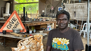 Person seated in a workshop beside a rusted metal object, with a weathered “GIVE WAY” road sign leaning on a workbench in the background and tools and materials scattered throughout the space.
