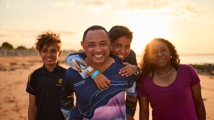 Four people standing close together on a sandy beach at sunset, facing the camera, with warm light behind them and the ocean and shoreline in the background.