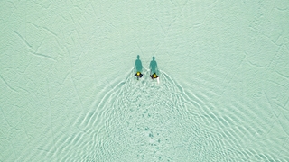 Two people wading through shallow, turquoise water, creating rippling patterns viewed from above.