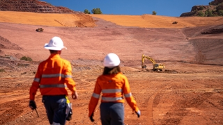 Two workers in high-visibility clothing and hard hats walk across a rehabilitated mine site, with earthmoving equipment operating in the background.