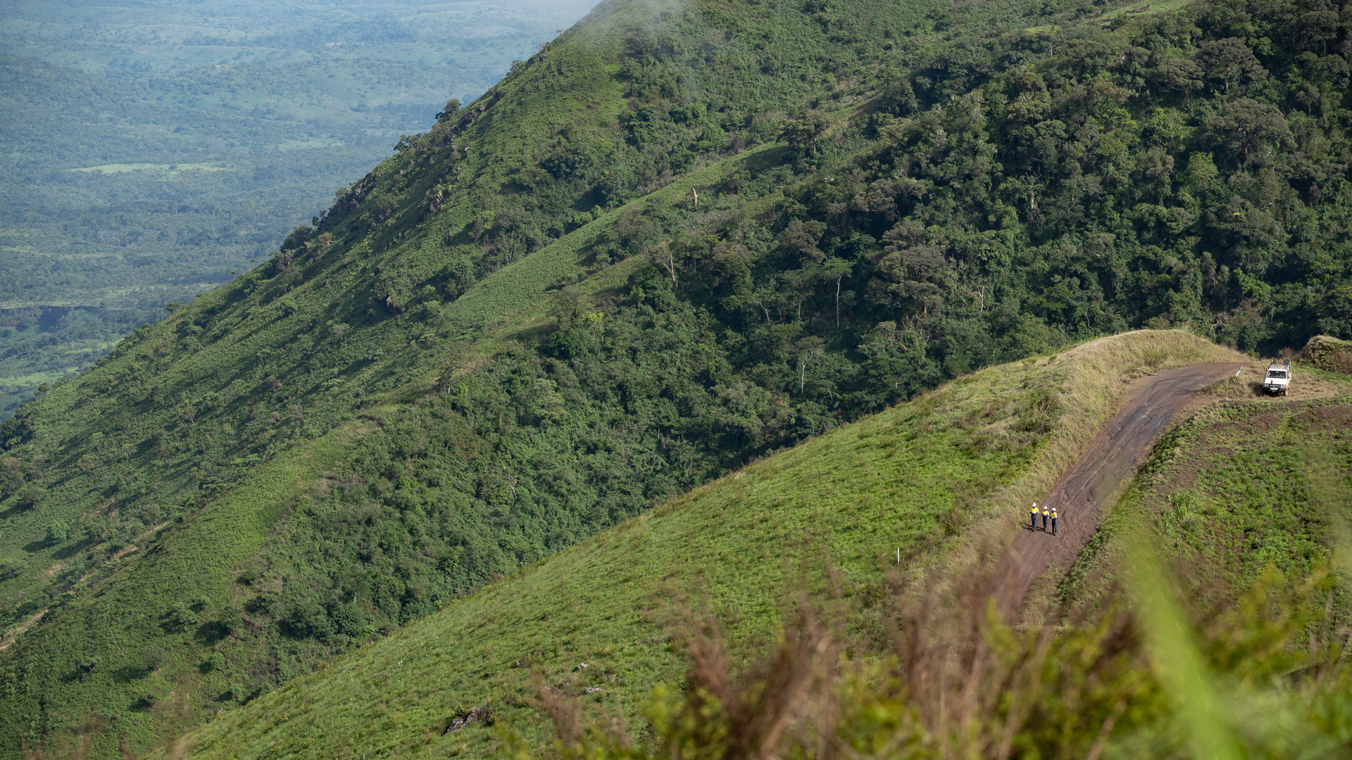 A wide view of lush green mountains and forested slopes, with a dirt road winding along a ridgeline. Three workers in high-visibility clothing walk on the road, while a white vehicle is parked further ahead overlooking the valley.