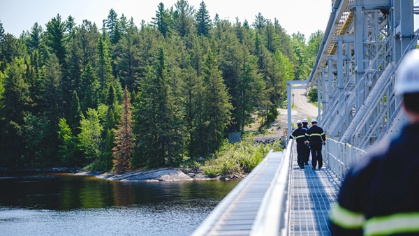 A group of workers wearing safety gear walk along a metal bridge structure over a body of water, with dense green forest in the background. The bridge has railings and appears to be part of an industrial facility. Another worker is visible in the foreground, partially out of focus.