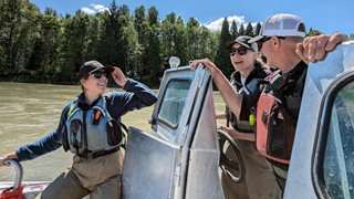 Three people wearing life jackets and caps stand close together on a small metal boat, talking near the steering console on a wide, muddy river. Forested trees line the riverbank in the background under a clear blue sky, and sunlight illuminates the scene.
