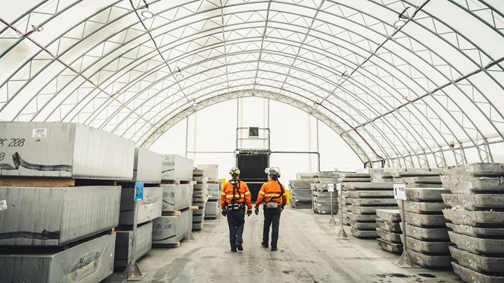 Two workers wearing high‑visibility safety clothing walk down the center aisle of a large industrial storage facility with a curved, translucent roof. Stacks of large aluminium or concrete blocks are arranged in rows on both sides