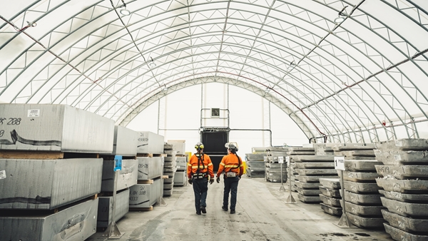 Two workers wearing high‑visibility safety clothing walk down the center aisle of a large industrial storage facility with a curved, translucent roof. Stacks of large aluminium or concrete blocks are arranged in rows on both sides