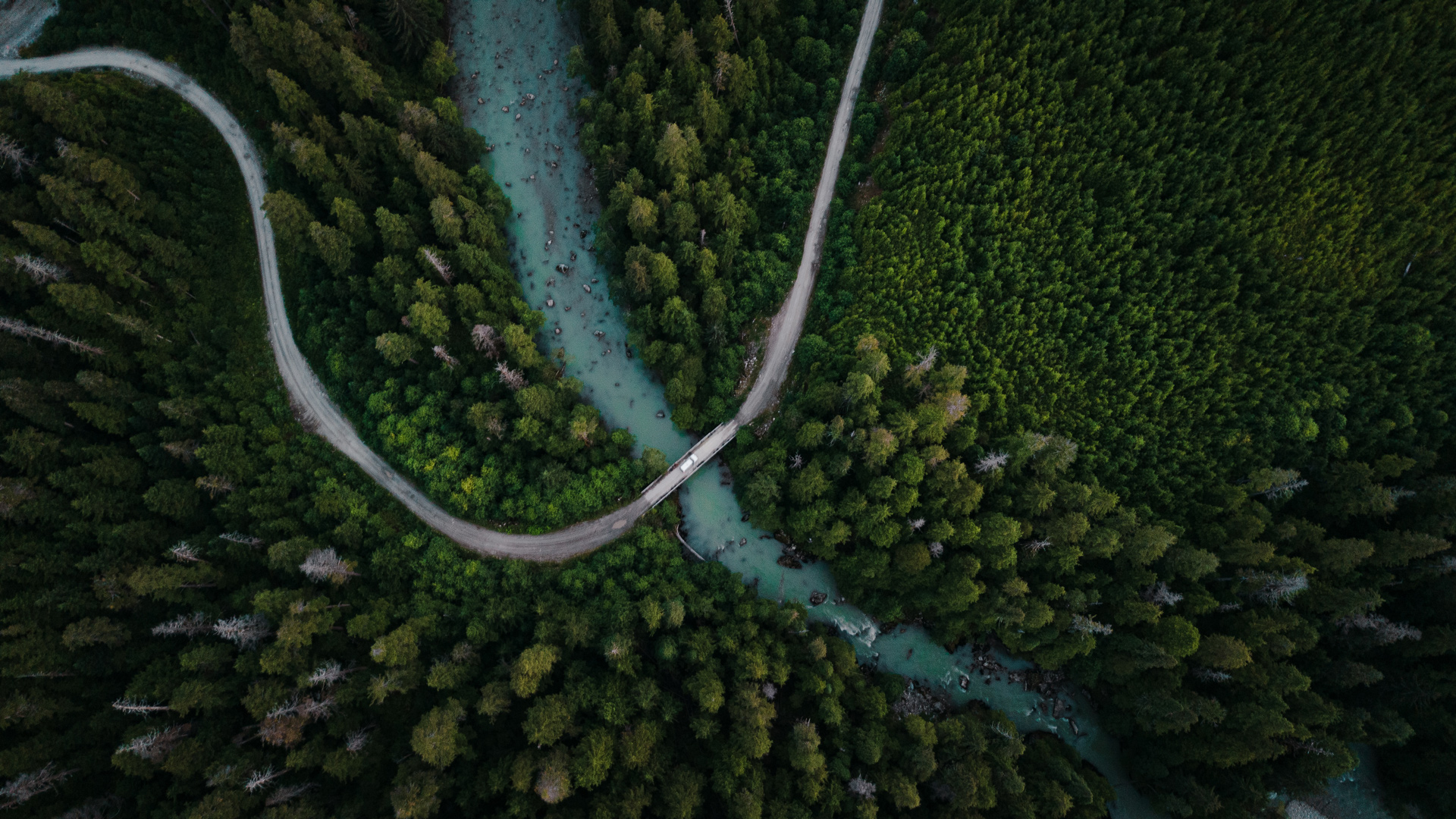 Aerial view of a winding road crossing a turquoise river through dense green forest.