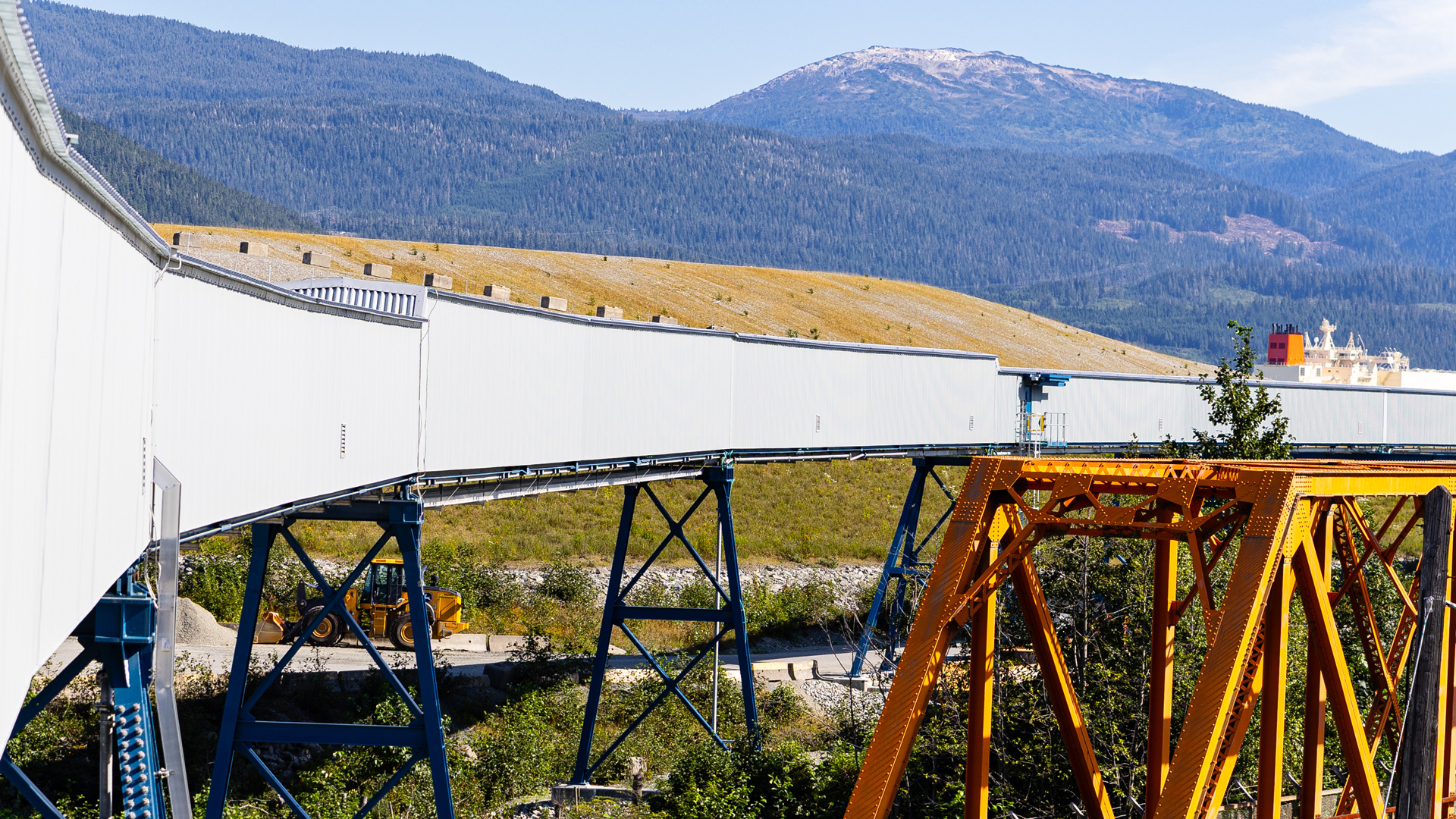 Elevated industrial conveyor belt supported by steel structures, with forested mountains in the background.