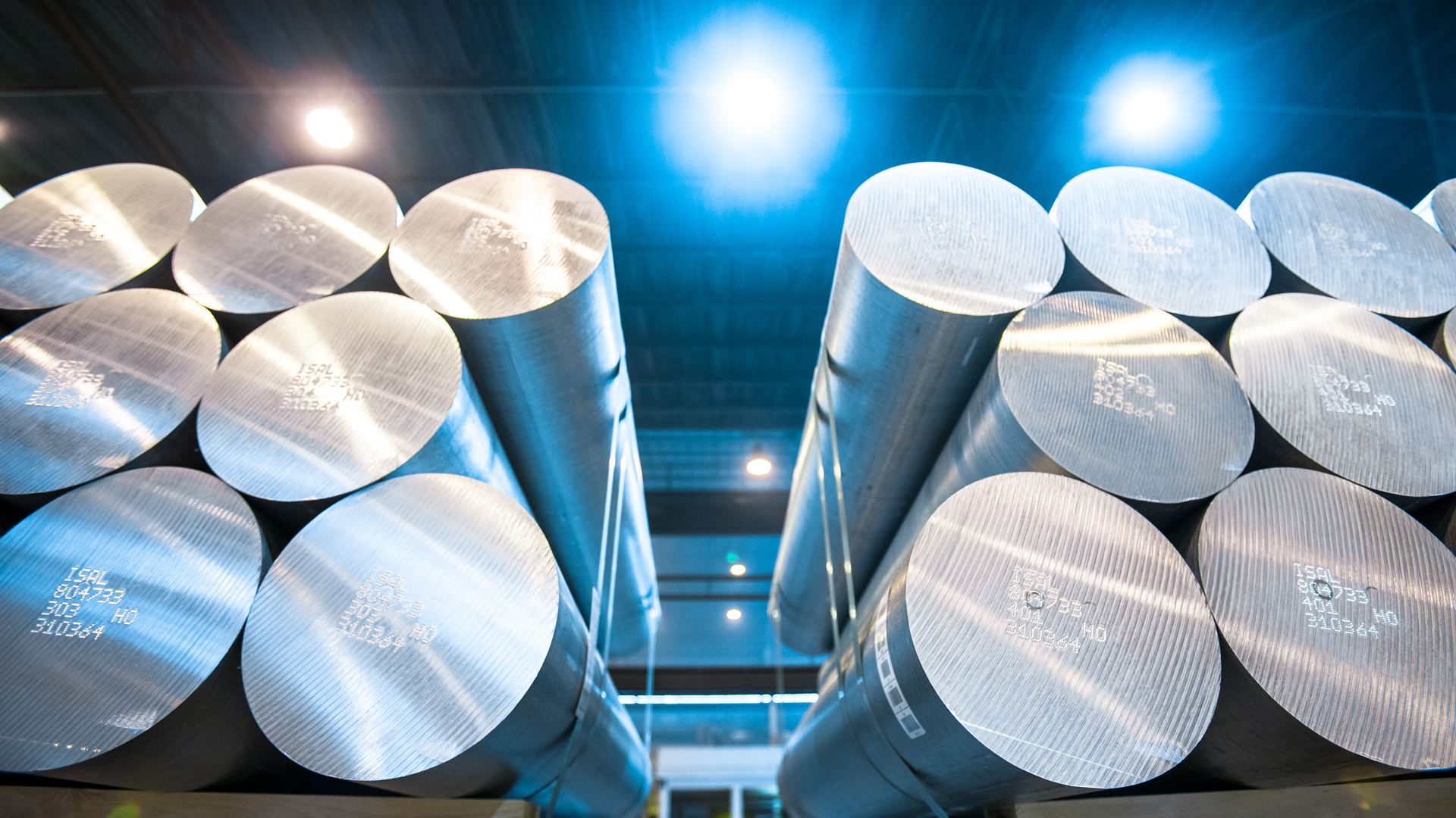 Stacked aluminium billets in a factory, illuminated by bright overhead lights