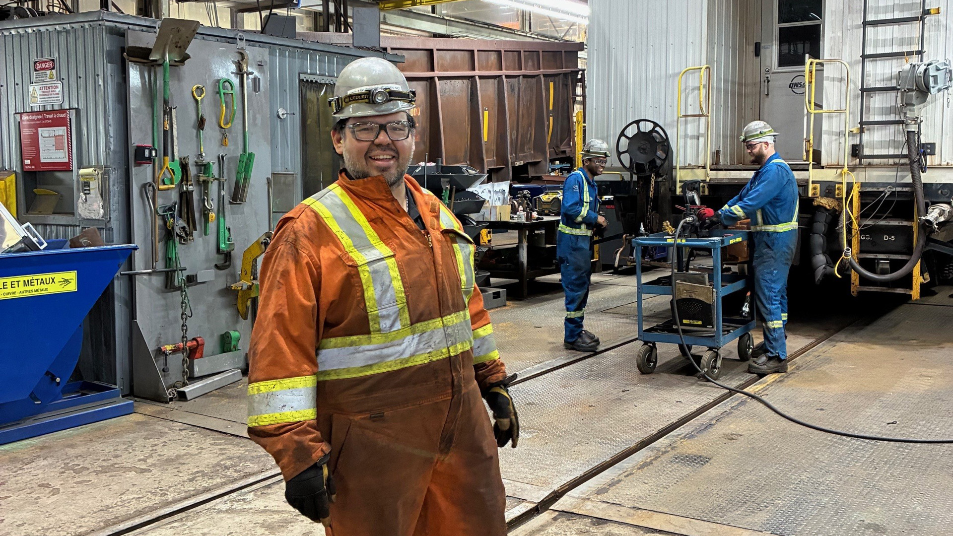 Industrial workshop interior with overhead steel beams and lighting. In the foreground, a worker wearing an orange coverall with reflective stripes, a hard hat, safety glasses, and gloves stands facing the camera.