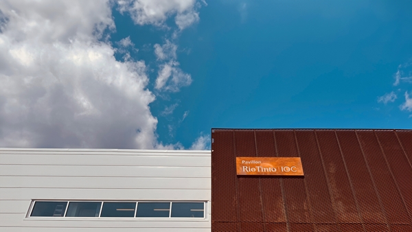 Modern building exterior with two contrasting facades—one white with narrow horizontal windows and one textured copper-toned panel—featuring a sign that reads “Pavillon Rio Tinto IOC” against a bright blue sky with scattered clouds.