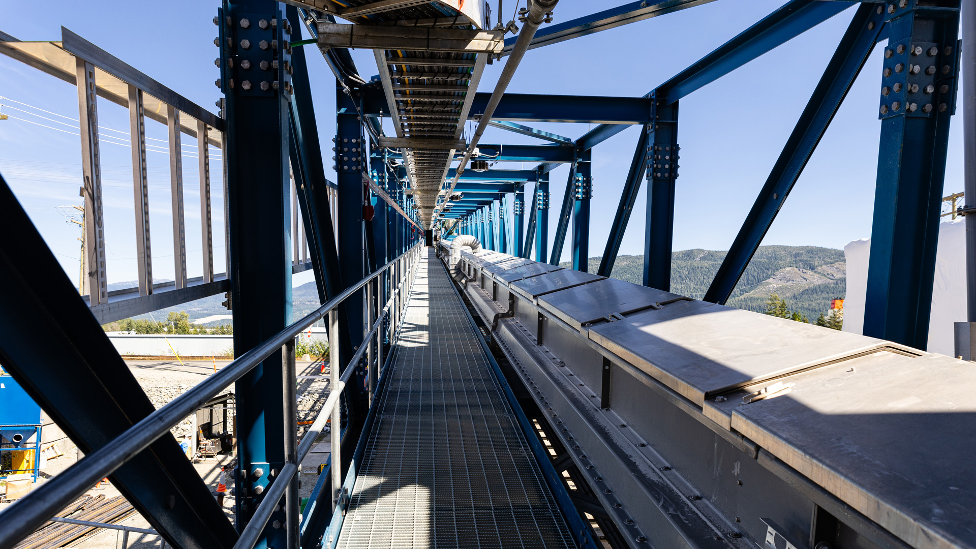 Inside view of an elevated industrial conveyor system, showing a long metal conveyor belt running alongside a grated walkway, enclosed by blue steel support beams, with hills visible in the distance under a clear sky