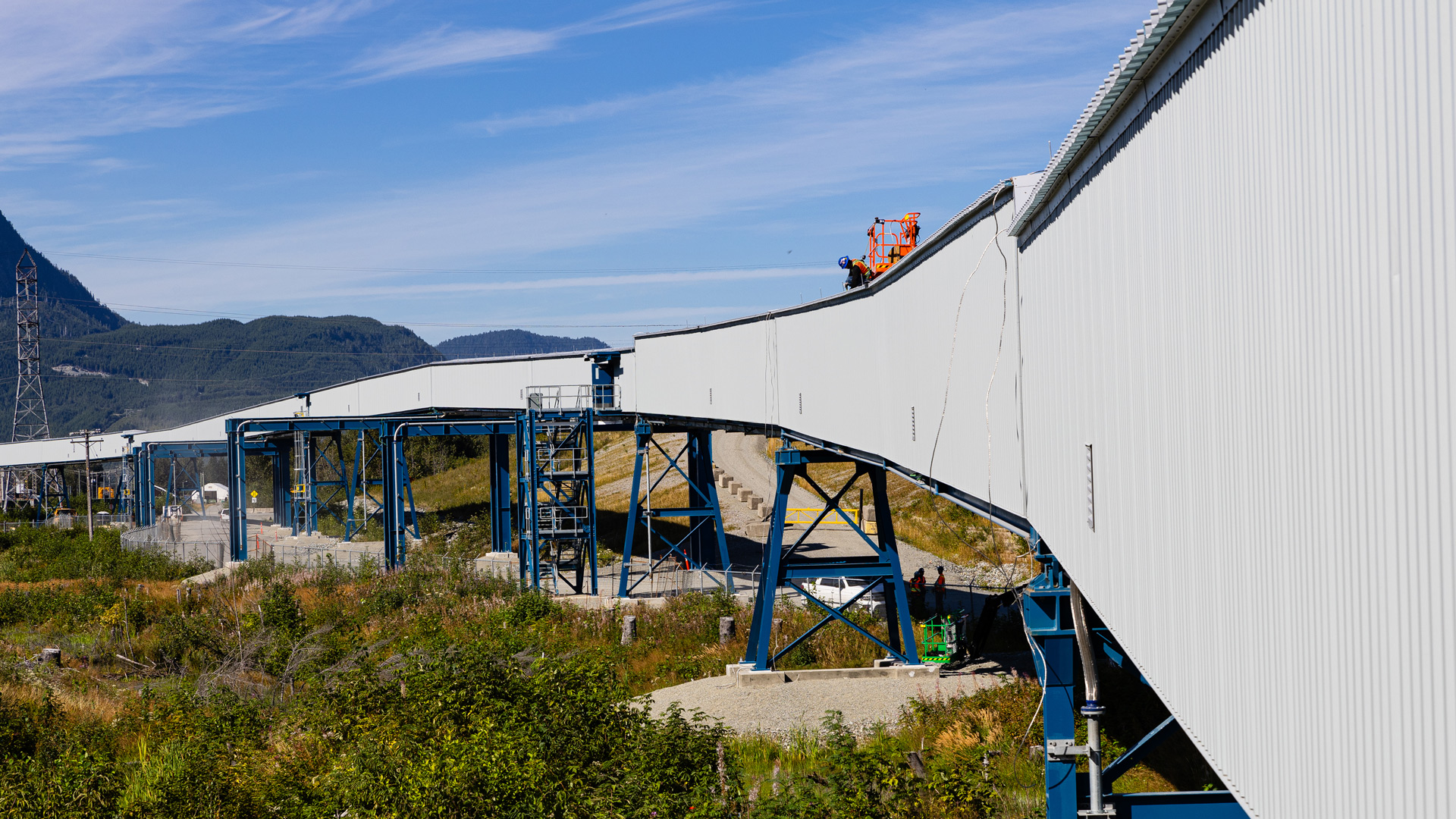 Long, elevated industrial conveyor structure supported by blue steel frames, extending across a grassy area with hills and mountains in the background under a clear blue sky.