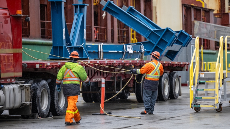 Workers in high‑visibility protective clothing guide large blue steel conveyor components being unloaded from a flatbed truck at an industrial facility, with safety barriers and equipment positioned nearby