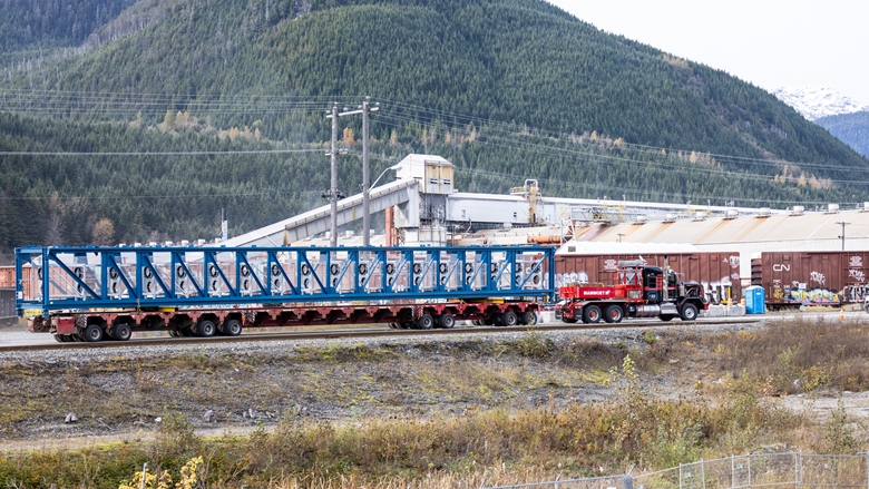 Long blue steel conveyor structure secured on a multi‑axle flatbed truck, parked at an industrial site with buildings, rail cars, and forested hills in the background.