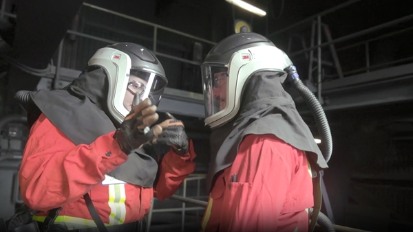 Two individuals in red protective suits and helmets with face shields and attached hoses, standing in an industrial setting with metal railings and equipment.