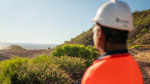 Person wearing a white safety helmet and bright orange high-visibility shirt looking out over a dry, rugged landscape with green shrubs and distant hills under a clear blue sky.